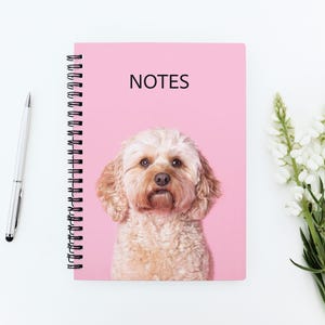 May include: A pink notebook with the word "NOTES" on the cover, featuring a dog's face. A silver pen is to the left, and white flowers are to the right. The notebook's cover has a light brown dog with a pink background.