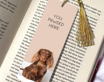 Peut inclure: Marque-page avec un chien teckel marron et le texte "YOU PAWSED HERE". Il est orné d'un pompon doré et placé dans un livre ouvert. Le chien a de longues oreilles.
