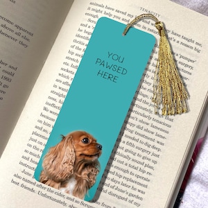 May include: A teal bookmark featuring the text "YOU PAWSED HERE" and a Cavalier King Charles Spaniel dog. It has a gold tassel and rests on an open book.
