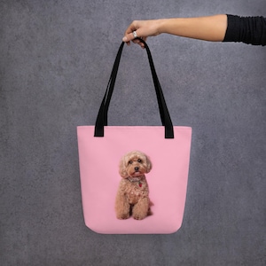 May include: A pink tote bag with a light brown, fluffy dog printed on the front. The dog has a red collar. The bag has black handles and is held up by a person's hand. The dog is centered on the bag.