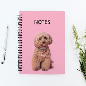 May include: Pink spiral-bound notebook with the word "NOTES" and a brown dog illustration. A silver pen and white flowers are next to the notebook on a white surface. The dog has a red strawberry charm.