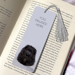 Op de afbeelding: Een boekenlegger met een zwarte dwergkeeshond puppy en de tekst "YOU PAWSED HERE." De boekenlegger heeft een zilveren kwast en ligt in een open boek. Een cadeau voor honden- en boekenliefhebbers.