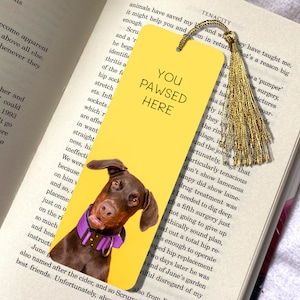 Może przedstawiać: Żółta zakładka z brązowym i czarnym psem rasy Doberman Pinscher, który wystaje z dołu. Zakładka zawiera tekst "You Pawsed Here" czarnymi literami.