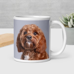 May include: White ceramic mug with a portrait of a Cavapoo dog, a cross between a Cavalier King Charles Spaniel and a Poodle. The dog has reddish-brown fur and a white patch on its chest. The background is grey.