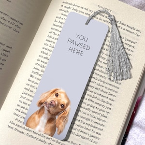 Könnte beinhalten: Ein hellblaues Lesezeichen mit dem Text "You Pawsed Here" und einem Bild eines braunen und weißen Hundes mit Schlappohren. Das Lesezeichen hat einen silbernen Quasten.