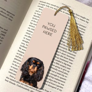 May include: A pink bookmark with a black and tan Cavalier King Charles Spaniel dog on it. The bookmark has the text "You Pawsed Here" in black.