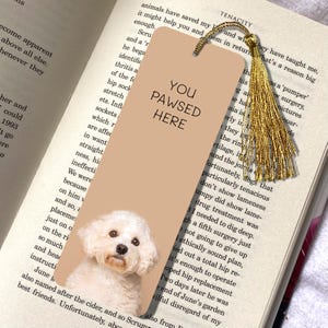 Op de afbeelding: Tan boekenlegger met een Bichon Frise hond en de tekst "YOU PAWSED HERE". Het heeft een gouden kwast en ligt in een boek. Een leuk cadeau voor hondenliefhebbers en boekenwurmen.