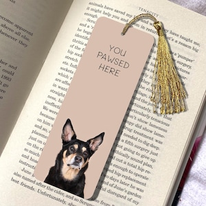 May include: A pink and brown bookmark with a black and white photo of a dog looking at the camera. The bookmark has the text "You Pawsed Here" in black text.