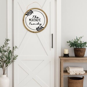 May include: A round wooden sign with the text "The McKenzie Family" in black script, surrounded by black leaf accents. The sign hangs on a white door with a black handle. A decorative shelf with plants and a candle is visible.