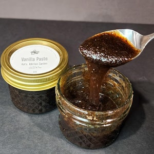 May include: Two glass jars filled with dark brown vanilla paste. The paste is being poured from one jar into the other using a silver spoon. The jar has a gold lid with a white label that reads "Vanilla Paste, Kat's Kitchen Garden".