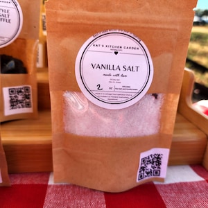 May include: A brown paper bag of vanilla salt with a clear window, labeled "Kat's Kitchen Garden." The label reads "Vanilla Salt" and "2 OZ." The bag is on a wooden display with a red and white checkered tablecloth.