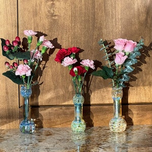 Könnte beinhalten: Drei Glasvasen mit einem farbenfrohen, geschwungenen Design halten rosa und rote Nelken. Die Vasen stehen auf einer braunen Arbeitsplatte.