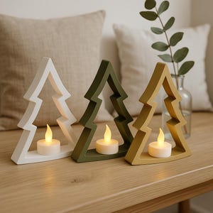 May include: Three tree-shaped candle holders in white, olive green, and mustard yellow. Each holder has a small, battery-operated candle inside, creating a warm glow. The candle holders are placed on a wooden surface.