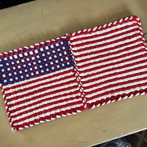 May include: Two woven American flag pot holders, one with a blue and white checkered pattern for the stars and the other with red and white stripes. The pot holders are joined together and have a white border.