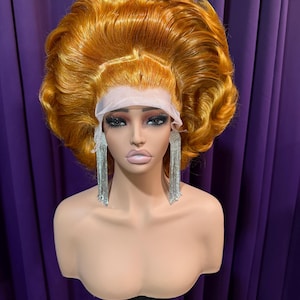 Può includere: Un busto di manichino che mostra una voluminosa parrucca arancione dorata con un frontale in pizzo. La parrucca è acconciata con riccioli grandi e morbidi. Il manichino indossa lunghi orecchini a nappa argentati. Lo sfondo è una tenda viola.