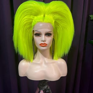 Könnte beinhalten: Ein Schaufensterpuppenkopf mit einer leuchtend neongrünen Perücke. Die Perücke hat einen voluminösen, gestuften Stil. Die Schaufensterpuppe hat ein komplettes Make-up, einschließlich kräftigem rotem Lippenstift und langen Wimpern. Schwarze Kettenohrringe sind zu sehen. Die Puppe steht auf einem Stativ.
