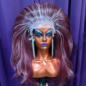 Könnte beinhalten: Ein Schaufensterpuppenkopf mit auffälligem Make-up und einer großen, voluminösen Perücke. Die Perücke hat burgunderrote und weiße Strähnen, die mit Höhe und Volumen gestylt sind. Lange, silberne Strass-Ohrringe hängen von den Ohren der Schaufensterpuuppe. Der Hintergrund ist ein violetter Vorhang.