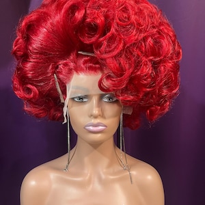 May include: A vibrant red wig with voluminous, curly hair. The wig is styled on a mannequin head, showcasing its full, textured appearance. The wig features a lace front and is designed to be worn. The mannequin has long, dangling earrings.