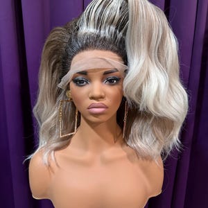 Może przedstawiać: Głowa manekina prezentująca blond i brązową perukę z wysokim kucykiem. Peruka ma falowane, sięgające do ramion włosy z koronkowym przodem. Manekin ma na sobie duże, kwadratowe złote kolczyki i pełny makijaż.