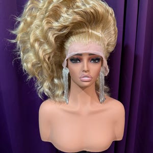 Può includere: Una parrucca bionda con capelli ondulati e voluminosi acconciati in una coda di cavallo alta. La parrucca è su una testa di manichino con un viso realistico e lunghi orecchini pendenti argentati. Lo sfondo è una tenda viola.