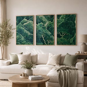 May include: Three framed landscape art prints showing a verdant mountain scene. The artwork hangs above a white sectional sofa with cushions and a wooden coffee table. A neutral rug and curtains complete the room's decor.