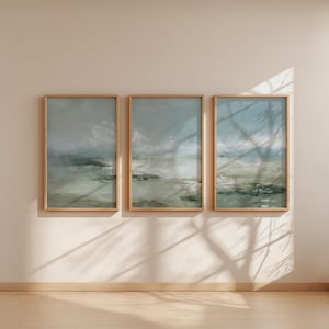 May include: Three framed landscape paintings with a muted color palette of blues, greens, and grays. The artwork is displayed on a beige wall, with natural light casting shadows across the scene. The frames are a light wood color.