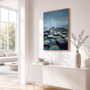 May include: A framed art print featuring a landscape of hexagonal rock formations leading to the sea under a cloudy sky. The artwork is displayed on a white wall above a white cabinet. A white vase with greenery is on the cabinet.