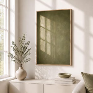 Könnte beinhalten: Ein vertikales, rechteckiges Wandbild mit einer olivgrünen, strukturierten Oberfläche in einem hellen Holzrahmen. Das Kunstwerk ist an einer Wand angebracht und fängt Licht- und Schattenmuster ein, die eine subtile visuelle Tiefe erzeugen.