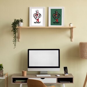 May include: Two framed illustrations of a person with a red and white design on a white background and a red and green design on a green background. The frames are white and the illustrations are titled "MKOF". The illustrations are on a wooden shelf above a desk with a computer, a lamp, and a potted plant.