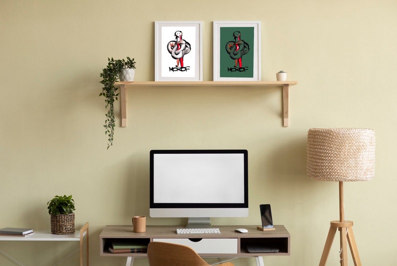 May include: Two framed illustrations of a person with a red and white design on a white background and a red and green design on a green background. The frames are white and the illustrations are titled "MKOF". The illustrations are on a wooden shelf above a desk with a computer, a lamp, and a potted plant.