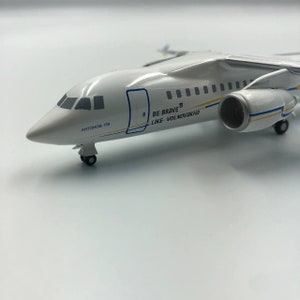 Puede incluir: Modelo de avión blanco con detalles en azul y amarillo. El texto "BE BRAVE LIKE VOLNOVAKHA" está impreso en el lateral del avión. El número de registro "АНТОНОВ-158" está impreso en la parte inferior del avión.
