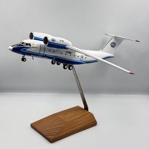 May include: A model airplane with a white and blue fuselage, two engines, and a silver stand. The model is mounted on a wooden base. The airplane has the text "D-36" and "MOTOR CIU" printed on it.