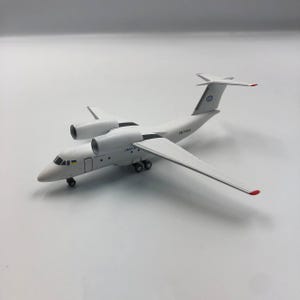 以下が含まれることがあります： 2つのジェットエンジン、翼、尾を持つ白い模型飛行機。飛行機は黒い車輪と、翼端と尾に赤いアクセントがあります。尾には「UR-74020」の文字があります。