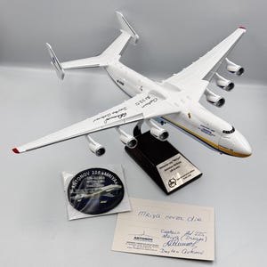 May include: A white model airplane of the Antonov An-225 Mriya cargo plane, with a black base and a small plaque that reads "Antonov An-225 'Mriya' Antonov Airlines Ukraine". The model is signed by the pilot, "Captain D.  Chernenko". There is a small round sticker with the words "Antonov 225 Mriya UR-82060 International Cargo Transporter" and a handwritten note that reads "Mriya never die Captain Al 225 Mriya (Dream) Daytro Antonov".