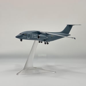 Könnte beinhalten: Ein graues Flugzeugmodell auf einem transparenten Kunststoffständer. Der Rumpf trägt den Schriftzug BE BRAVE LIKE HOSTOMEL und eine kleine ukrainische Flagge nahe dem Cockpit. Das Flugzeug hat zwei Strahltriebwerke unter den Flügeln.