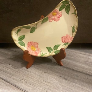 May include: An off-white ceramic serving dish with a floral design. The dish features pink roses and green leaves, with a gold trim. It is displayed on a brown wooden stand, showcasing its unique shape and design.