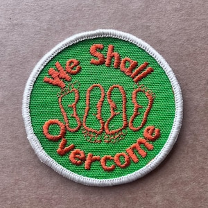 May include: Embroidered patch with the words "We Shall Overcome" in orange thread on a green background. The patch has a white border and features three sets of footprints.