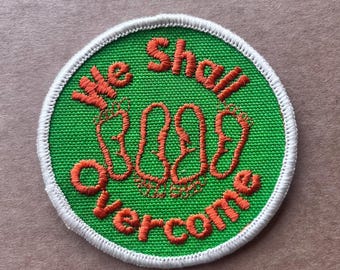 May include: Embroidered patch with the words "We Shall Overcome" in orange thread on a green background. The patch has a white border and features three sets of footprints.