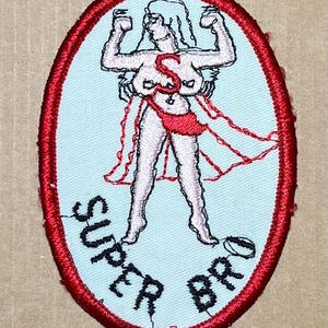 May include: An oval embroidered patch with a light blue background and a thick red border. It features a central figure with arms raised, wearing a red cape and a red letter S on the chest. The black text SUPER BRO is stitched below the figure.