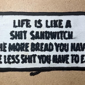 May include: A rectangular embroidered patch with a white background and a thick black border. The patch features black embroidered text that reads: LIFE IS LIKE A SHIT SANDWITCH THE MORE BREAD YOU HAVE THE LESS SHIT YOU HAVE TO EAT.