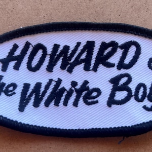 May include: Oval-shaped embroidered patch with the text "HOWARD & the White Boys" in black script on a white background. The patch has a black border.