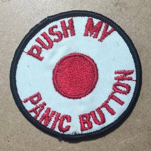 May include: A circular embroidered patch with a white background and a black border. The center features a solid red circle. The red embroidered text PUSH MY PANIC BUTTON is arranged in a circular pattern around the central red circle.