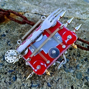 Puede incluir: Una pieza de arte de ensamblaje única y hecha a mano. Presenta una base rectangular roja adornada con un tenedor plateado, resortes, discos metálicos y puntas doradas. La obra de arte se encuentra sobre un fondo gris texturizado.