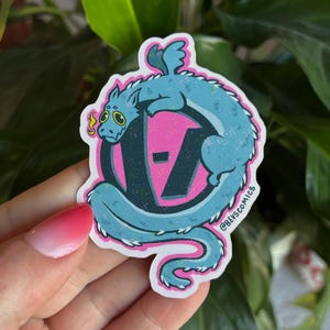 Peut inclure: Autocollant représentant un dragon turquoise enroulé autour d'un logo rose et noir. Le dragon a des accents jaunes et l'autocollant a une bordure rose. Le texte "@BEVSCOMICS" est imprimé sur l'autocollant.