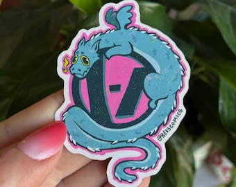 Op de afbeelding: Een sticker met een turquoise draak die zich om een roze en zwart logo wikkelt. De draak heeft gele accenten en de sticker heeft een roze rand. De tekst "@BEVSCOMICS" staat op de sticker.
