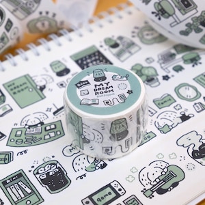 May include: A roll of decorative washi tape with a repeating pattern of green and black illustrations of a room, including a lamp, book, and cat. The tape roll has the text "MY DREAM ROOM" on it. The tape is on a white spiral notebook.