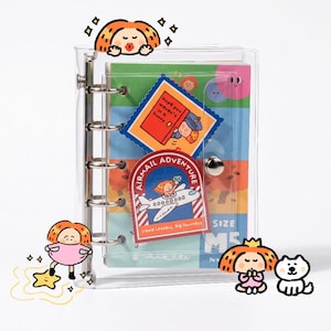 May include: A clear plastic M5 binder with a metal ring mechanism. The cover features colorful stickers including one that says "Hope you weren't in a hurry" and another labeled "AIRMAIL ADVENTURE" with a cartoon airplane and character.