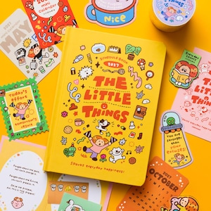 May include: A yellow 2027 schedule book titled The Little Things surrounded by various stationery items including a mini calendar, stickers, a roll of washi tape, and cards featuring colorful cartoon illustrations and motivational text.