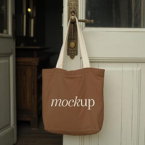 May include: A brown tote bag with white straps hangs from a vintage door handle. The word "mockup" is printed in white script on the front of the bag. The door is painted a light green color.
