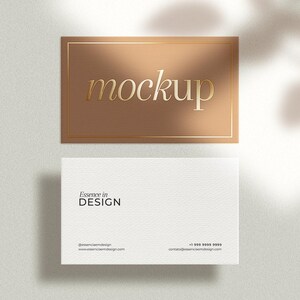 May include: A mockup business card design featuring a gold-colored rectangular card with the word "mockup" in gold lettering. Below is a white business card with the text "Essence in DESIGN" and contact information. The design is set against a neutral background.